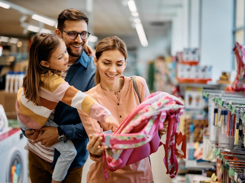 Happy little girl pointing at backpack while buying school supplies with her parents in supermarket.