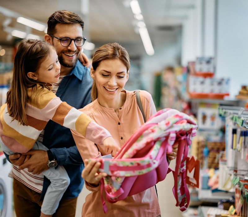 Happy little girl pointing at backpack while buying school supplies with her parents in supermarket.